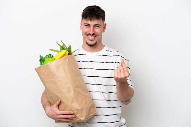 Młody kaukaski mężczyzna trzyma torbę na zakupy spożywcze na białym tle robiąc gest pieniędzy