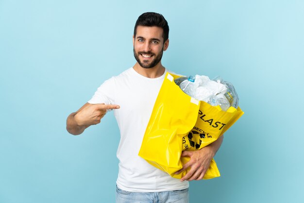 Młody kaukaski mężczyzna trzyma torbę pełną plastikowych butelek do recyklingu na białym tle na niebieskiej ścianie i wskazuje ją