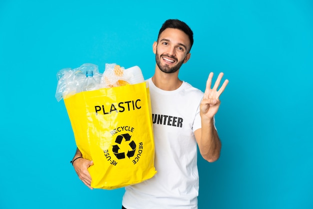 Młody kaukaski mężczyzna trzyma torbę pełną plastikowych butelek do recyklingu na białym tle na niebieskim tle szczęśliwy i liczy trzy palcami