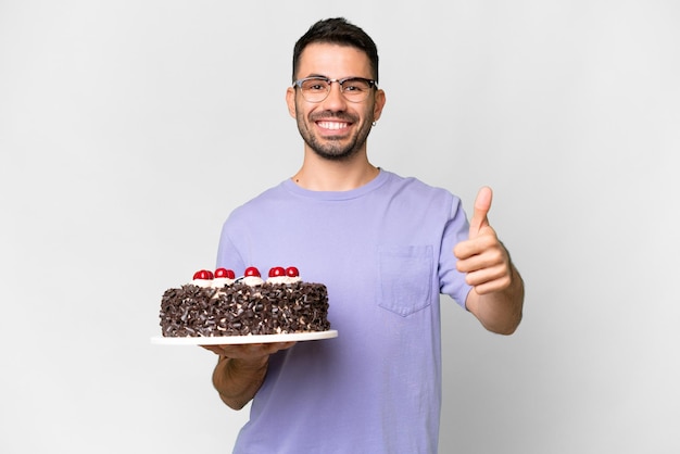 Młody kaukaski mężczyzna trzyma tort urodzinowy odizolowany na białym tle z kciukami do góry, ponieważ stało się coś dobrego