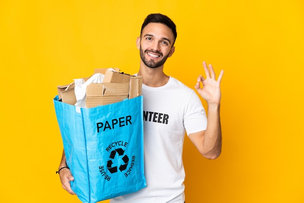 Młody kaukaski mężczyzna trzyma worek recyklingu pełen papieru do recyklingu na białym tle na białym tle Wyświetlono znak ok palcami