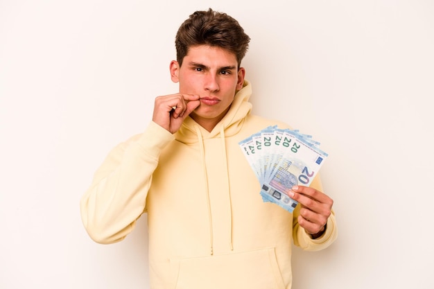 Młody kaukaski mężczyzna trzymający banknoty na białym tle z palcami na ustach zachowujący tajemnicę