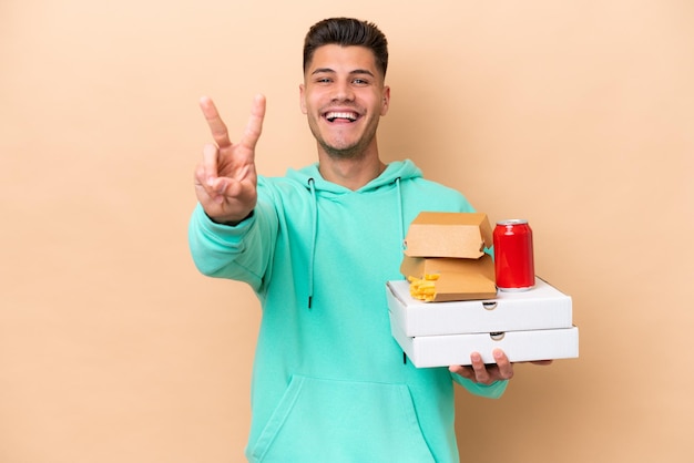 Młody kaukaski mężczyzna trzymający fast food na beżowym tle uśmiechający się i pokazujący znak zwycięstwa