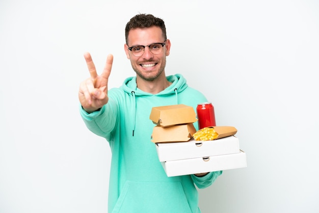 Młody kaukaski mężczyzna trzymający fast food na białym tle uśmiechający się i pokazujący znak zwycięstwa