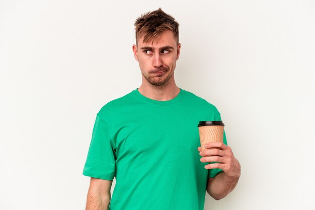 Zdjęcie młody kaukaski mężczyzna trzymający kawę na wynos na białym tle zdezorientowany, czuje się niepewny i niepewny.