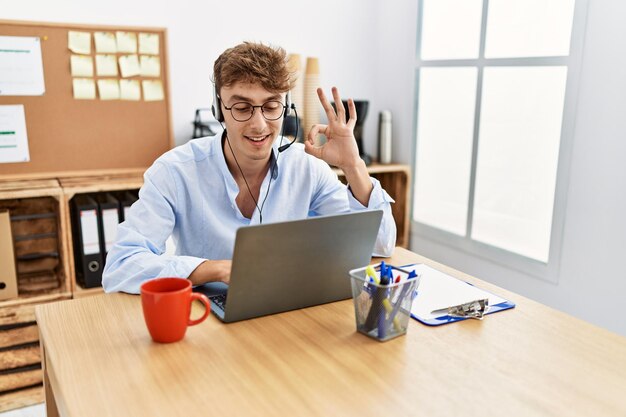 Młody kaukaski mężczyzna ubrany w zestaw słuchawkowy agenta call center w biurze robi znak ok palcami, uśmiechając się przyjaźnie gestykulując doskonały symbol