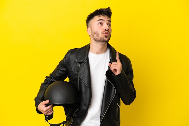 Młody kaukaski mężczyzna w kasku motocyklowym na żółtym tle myśli o pomyśle wskazującym palec w górę