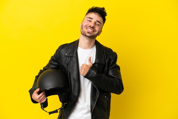Młody kaukaski mężczyzna z kaskiem motocyklowym na żółtym tle dumny i zadowolony z siebie