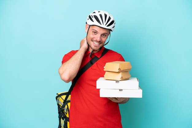 Młody Kaukaski Mężczyzna Z Plecakiem Termicznym I Trzymający Fast Food Odizolowany Na Niebieskim Tle śmiejąc Się