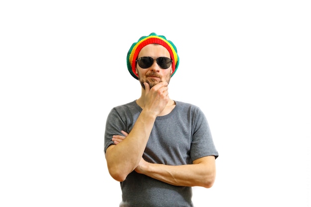 Młody kaukaski myśli człowiek w kapeluszu rasta, okularach przeciwsłonecznych i szarym t-shirt na białym tle.