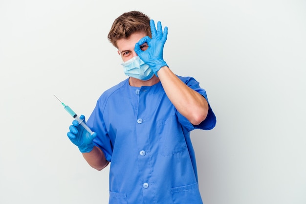 Młody kaukaski pielęgniarka mężczyzna przygotowany do podania szczepionki na białym tle na białej ścianie podekscytowany utrzymując ok gest na oko.