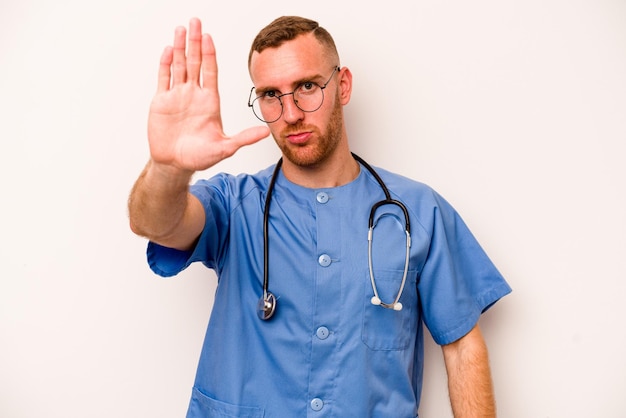 Młody kaukaski pielęgniarz mężczyzna na białym tle stojący z wyciągniętą ręką pokazując znak stopu, zapobiegający