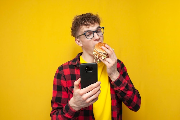 młody kędzierzawy facet w okularach trzyma dużego burgera i używa smartfona, student je fast food