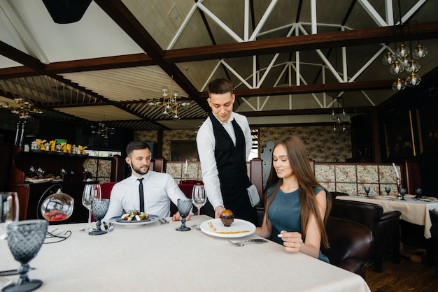 Młody kelner w stylowym fartuchu serwuje stolik z piękną parą w wytwornej restauracji. Obsługa klienta.