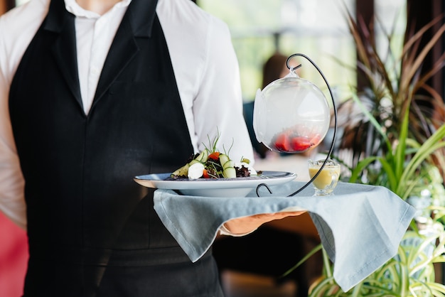 Młody kelner w stylowym mundurze stoi z wykwintnym daniem na tacy przy stole w pięknym zbliżeniu restauracji. Działalność restauracyjna na najwyższym poziomie.