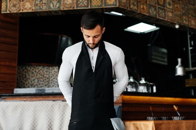 Młody kelner z brodą zakłada fartuch i przygotowuje się do pracy w eleganckiej restauracji