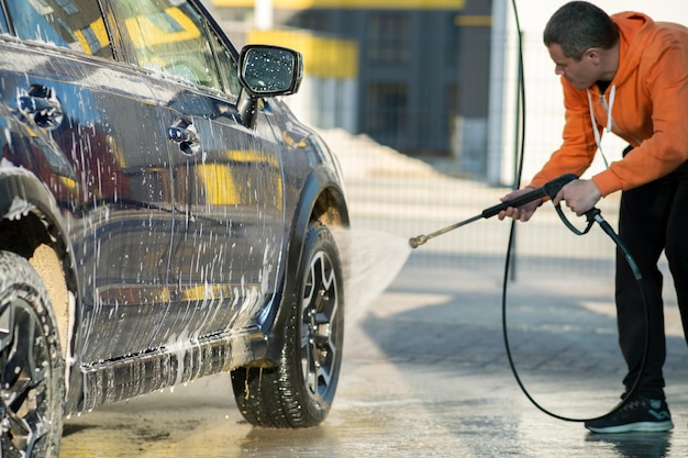 Młody kierowca człowiek mycie samochodu bezdotykowym strumieniem wody pod wysokim ciśnieniem w samoobsługowej myjni samochodowej.