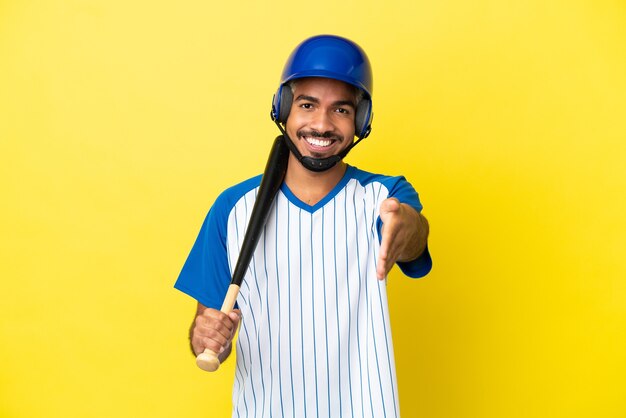 Młody kolumbijski latynoski mężczyzna grający w baseball na żółtym tle, ściskając ręce, aby zamknąć dobrą ofertę