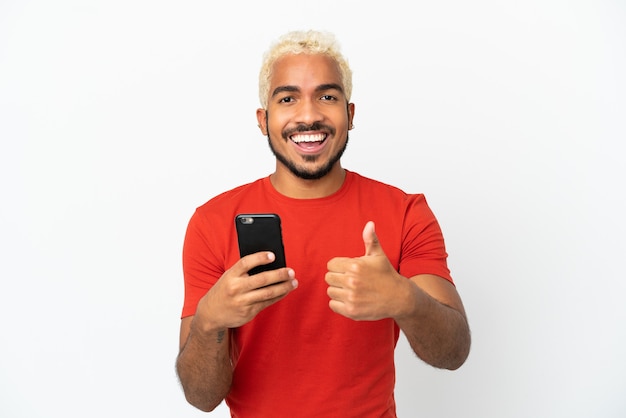Młody kolumbijski przystojny mężczyzna na białym tle przy użyciu telefonu komórkowego podczas robienia kciuków w górę