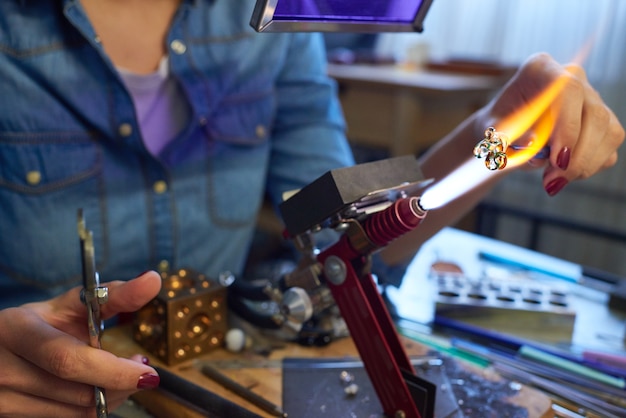 Młody kreatywny żeński mistrz lampworking siedzi przy stole przed palnikiem i przetwarza szklany przedmiot podczas spalania go w strumieniu ognia