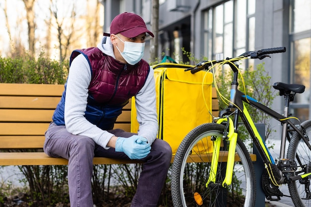 Młody kurier w masce medycznej dostarczający jedzenie z żółtym plecakiem termicznym, jeżdżący na rowerze po mieście. Koncepcja usługi dostawy żywności.