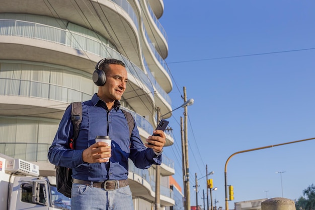 Młody łaciński mężczyzna z słuchawkami patrzący na swój telefon komórkowy i trzymający papierowy kubek z kawą