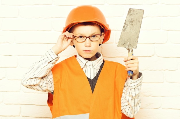 Zdjęcie młody ładny konstruktor chłopiec w pomarańczowym mundurze i kasku lub kasku w okularach, trzymając narzędzie lub szpachlę na białym tle ściany z cegły