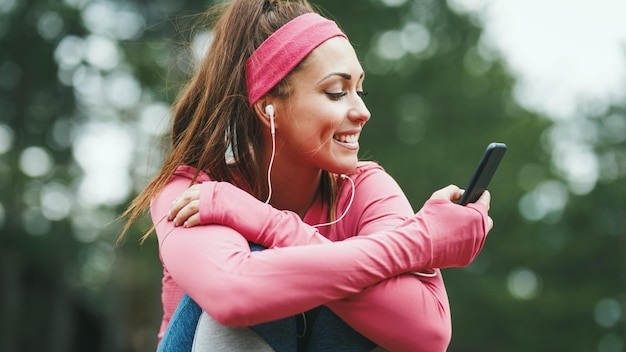 Młody ładny szczęśliwy biegaczka słuchania muzyki, przy użyciu smartfona i przerwy po joggingu w słonecznym lesie.