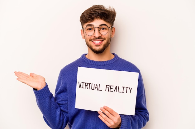 Młody latynoski mężczyzna trzymający afisz wirtualnej rzeczywistości na białym tle pokazujący miejsce na dłoni i trzymający inną rękę w talii