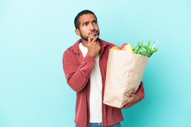 Młody latynoski mężczyzna trzymający torbę na zakupy spożywcze na białym tle na niebieskim tle i patrzący w górę