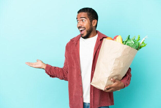 Młody latynoski mężczyzna trzymający torbę na zakupy spożywcze na białym tle na niebieskim tle z wyrazem zaskoczenia, patrząc z boku