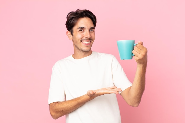 Młody latynoski mężczyzna uśmiecha się radośnie, czuje się szczęśliwy, pokazuje koncepcję i trzyma kubek z kawą