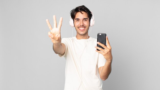 Młody latynoski mężczyzna uśmiechający się i wyglądający przyjaźnie, pokazujący numer trzy ze słuchawkami i smartfonem