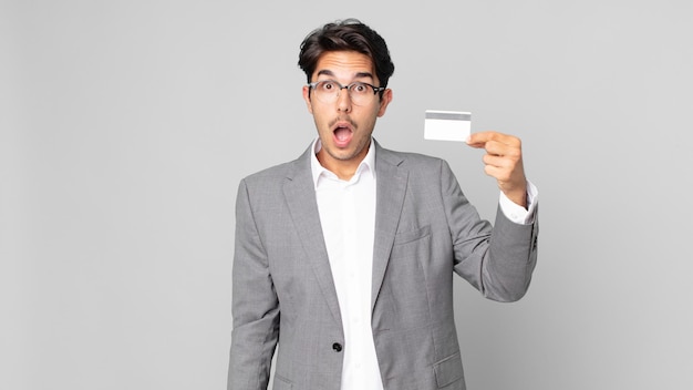 Młody latynoski mężczyzna wyglądający na bardzo zszokowanego lub zdziwionego i trzymający kartę kredytową