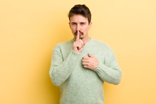 Zdjęcie młody latynoski mężczyzna wyglądający poważnie i skrzyżowany z palcem przyciśniętym do ust, domagający się ciszy lub spokoju dochowania tajemnicy