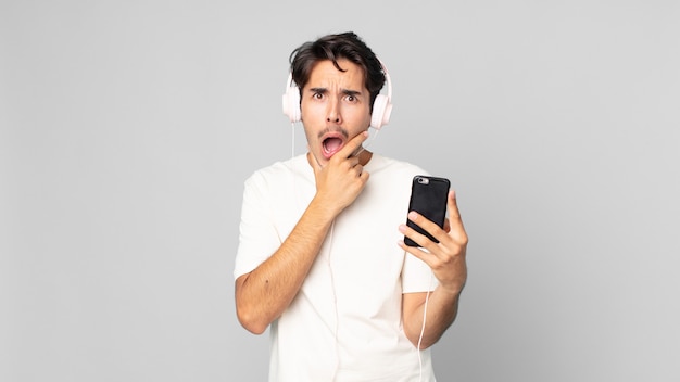 Młody latynoski mężczyzna z szeroko otwartymi ustami i oczami oraz ręką na brodzie ze słuchawkami i smartfonem