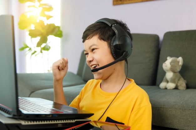 Młody Mały śliczny Azjatycki Chłopiec Z Lat 10 W Słuchawkach Siedzi W Domu W Salonie, Używając Laptopa Do Nauki Na Odległość Online I Podnosi Pięść Z Podekscytowanym I Pewnym Siebie