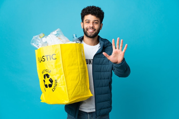 Młody marokańczyk trzymający torbę pełną plastikowych butelek do recyklingu, licząc palcami pięć