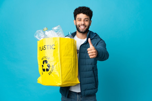 Młody Marokańczyk trzymający torbę pełną plastikowych butelek do recyklingu na odizolowanej ścianie z kciukami do góry, ponieważ wydarzyło się coś dobrego