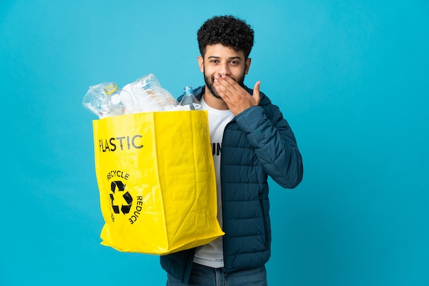 Młody marokański mężczyzna trzyma torbę pełną plastikowych butelek do recyklingu na odizolowanych szczęśliwych i uśmiechniętych zakrywających usta ręką