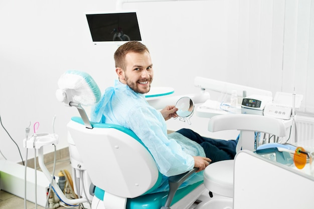 Młody męski klient uśmiecha się do kamery podczas sprawdzania zębów w lustrze w nowoczesnej białej gabinecie dentysty.