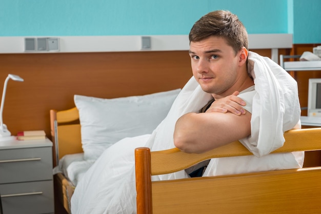 Młody mężczyzna chory opierając się na łożu szpitalnym przykrytym kołdrą na oddziale szpitalnym. Koncepcja opieki zdrowotnej