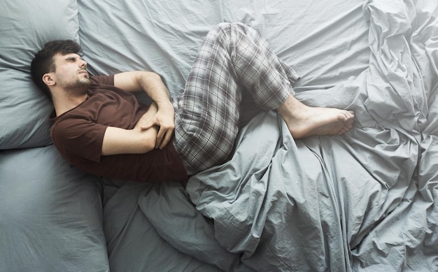 Młody mężczyzna cierpiący na ból brzucha, leżący w łóżku w domu, widok z góry. Koncepcja problemów zdrowotnych