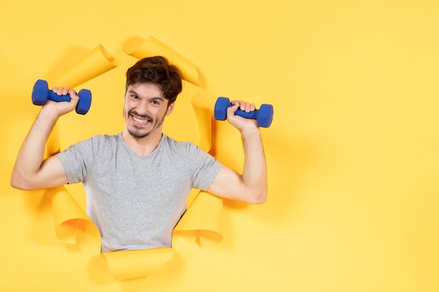 młody mężczyzna ćwiczący z niebieskimi hantlami na żółtym tle ćwiczenia sportowe siłownia