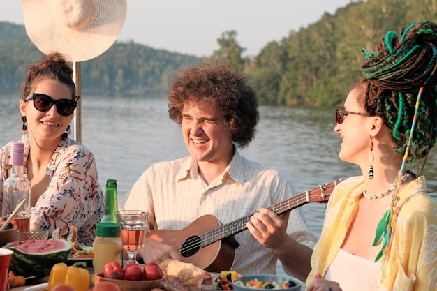 Młody mężczyzna gra na gitarze z dwiema kobietami siedzącymi między nim i śpiewającymi podczas lunchu na łonie natury