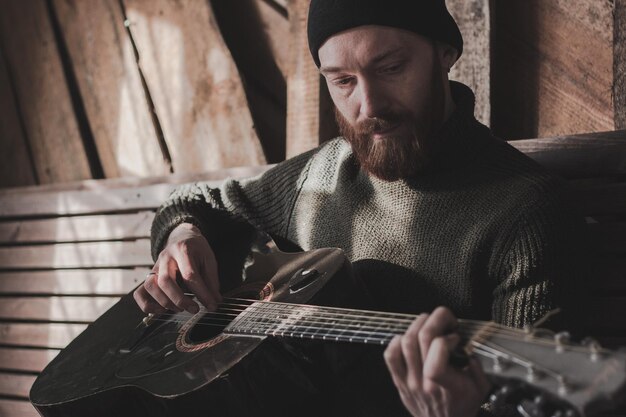 Młody mężczyzna grający na gitarze siedząc przy ścianie.