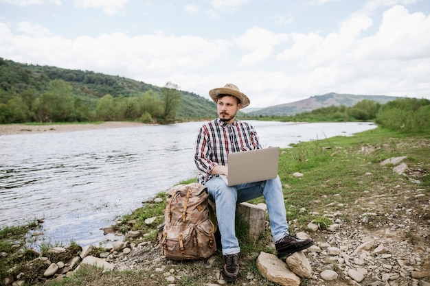 Młody mężczyzna hipster bloger pracuje zdalnie na laptopie na świeżym powietrzu podczas podróży w góry