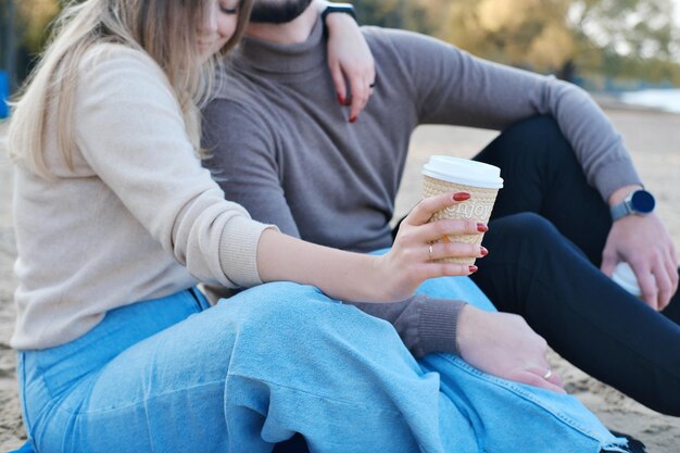 Młody mężczyzna i kobieta siedzą nad brzegiem jeziora, pijąc kawę i przytulając Skoncentruj się na szkle z kawą