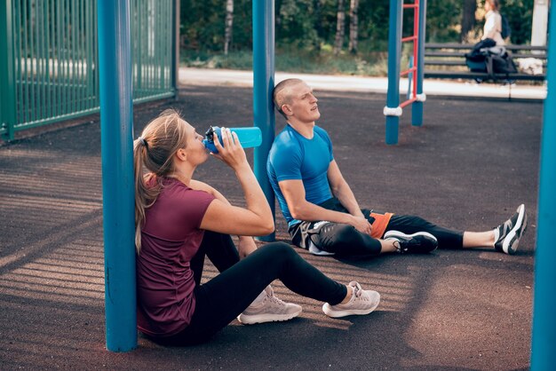 młody mężczyzna i kobieta sportowcy siedzą na placu zabaw i piją po wotkoucie