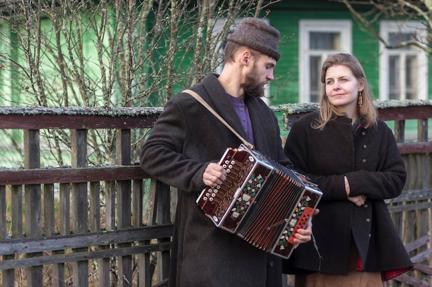 Zdjęcie młody mężczyzna i kobieta w rosyjskich ubraniach spacerują po wiosce i grają na akordeonie